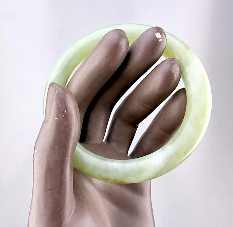 May include: A pale green jade bangle bracelet. The smooth, circular bracelet is held by a hand. The jade has a translucent quality, with variations in color. The bracelet is a classic piece of jewelry.