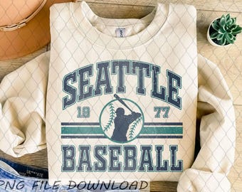 May include: Cream-colored sweatshirt featuring the text "SEATTLE BASEBALL" in a distressed, navy blue font. A baseball player silhouette is in a circle with the numbers "19" and "77". The sweatshirt has a crew neck.