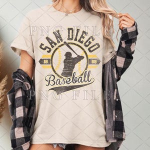 May include: Cream-colored t-shirt featuring a distressed San Diego Baseball logo with a batter silhouette, baseball, and the years 1969 and 1989. The shirt has a vintage aesthetic.