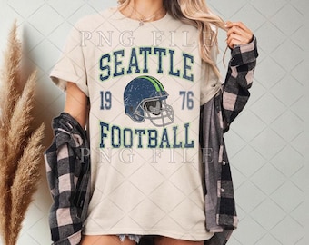 May include: Cream-colored t-shirt with the words "SEATTLE FOOTBALL" in blue and green, along with a football helmet graphic. The numbers "19" and "76" are also displayed. The shirt has a vintage, distressed look.