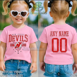Peut inclure: Un t-shirt rose clair à manches courtes arborant le logo des New Jersey Devils sur le devant avec le texte New Jersey Devils Est 1974. Le dos du t-shirt affiche le texte Any Name au-dessus du numéro 00 dans une police athlétique rouge et noire.