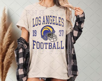 Op de afbeelding: Crèmekleurig T-shirt met de blauwe tekst "LOS ANGELES FOOTBALL", een blauw-gele footballhelm-afbeelding en de cijfers "19" en "37". Het shirt wordt gedragen met een zwart-grijs geruit overhemd.