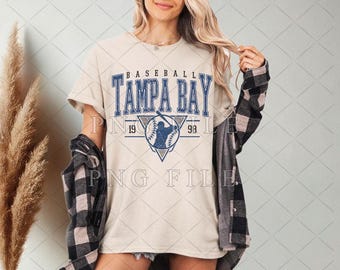 Pode incluir: Uma camiseta bege com um gráfico de beisebol azul e branco e o texto "BASEBALL TAMPA BAY 19 99".