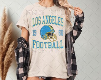 Op de afbeelding: Crèmekleurig T-shirt met de tekst "LOS ANGELES FOOTBALL 1960" in blauw. Een blauwe voetbalhelm met een gele streep staat in het midden. Het shirt wordt gedragen met een zwart-grijs geruit overhemd.