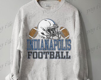 Peut inclure: Un sweat-shirt gris clair avec un casque de football américain et deux ballons. Le texte "INDIANAPOLIS FOOTBALL" est imprimé en bleu et gris. Le sweat-shirt est suspendu à un cintre en bois.