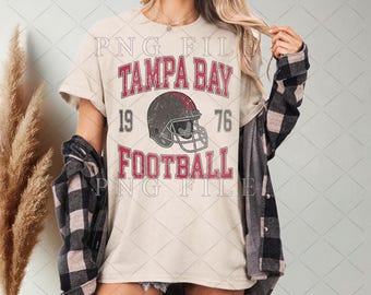 Pode incluir: Camiseta creme com o texto "TAMPA BAY FOOTBALL" em vermelho, e um gráfico de capacete de futebol desgastado. Os números "19" e "76" são impressos acima e abaixo do capacete. A camiseta faz parte de uma coleção de roupas esportivas.