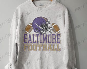 Può includere: Una felpa grigio chiaro con un casco da football viola e due palloni da football. Il testo "BALTIMORE FOOTBALL" è stampato in viola e oro. La felpa è appesa a una gruccia di legno.