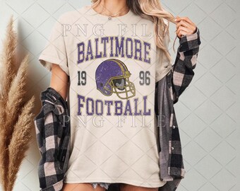 Può includere: T-shirt color crema con la scritta "BALTIMORE FOOTBALL 1996" in viola. Il design include un casco da football viola. La maglietta è indossata con una camicia a quadri nera e grigia.
