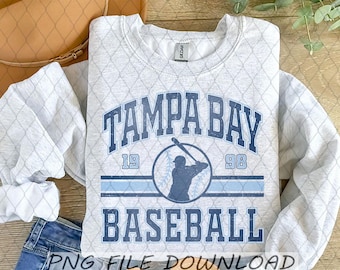 Pode incluir: Um moletom cinza claro com um padrão de losangos e as palavras "TAMPA BAY BASEBALL" em azul. O design inclui uma silhueta de jogador de beisebol e os números "19" e "98". Uma bolsa marrom e jeans azuis são visíveis.