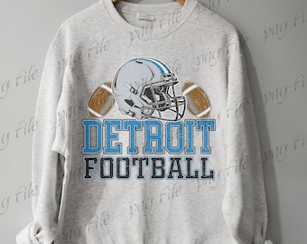Könnte beinhalten: Hellgraues Sweatshirt mit Footballhelm und zwei Fußbällen. Der Text "DETROIT FOOTBALL" ist in Blau und Schwarz gedruckt. Das Sweatshirt hängt an einem Holzkleiderbügel.