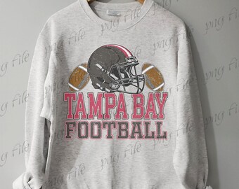 Pode incluir: Camisola cinza claro com um capacete de futebol americano e duas bolas. O texto "TAMPA BAY FOOTBALL" está impresso em rosa e cinza. A camisola está pendurada num cabide de madeira.