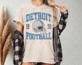 Könnte beinhalten: Beiges T-Shirt mit einem blau-grauen Footballhelm-Motiv. Das Shirt zeigt den Text "DETROIT FOOTBALL 1930" in Blau, mit einem Vintage-Look. Das Shirt wird mit einem schwarz-grauen karierten Hemd getragen.