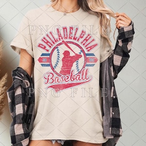 Könnte beinhalten: Beiges T-Shirt mit einem verblassten Grafikdesign eines Baseballs, einer Schlagmann-Silhouette und den Worten "Philadelphia Baseball". Das Design verwendet rote und blaue Akzente, mit dem Text "18" und "03" auf beiden Seiten des Baseballs.