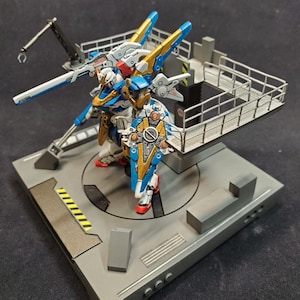May include: A blue, gold, and white robot action figure with a large shield and a gun, standing on a gray platform with a metal railing.