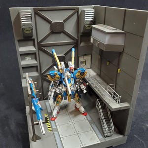 May include: A detailed model of a blue, white, and gold mecha robot standing in a gray diorama. The robot has intricate detailing and red accents. The diorama includes a metal staircase, a platform, and industrial-style walls.