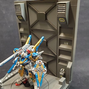 Puede incluir: Una figura de acción de robot azul, dorado y blanco de pie en una plataforma gris con un fondo gris y negro. El robot sostiene un arma grande de plata.