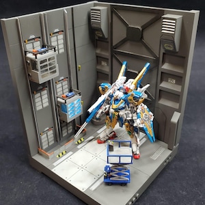 May include: A detailed model of a futuristic robot warrior, predominantly white, blue, and gold, stands in a gray, industrial-themed diorama. The scene includes miniature elevators, a small blue lift, and a textured floor, creating a dynamic display.