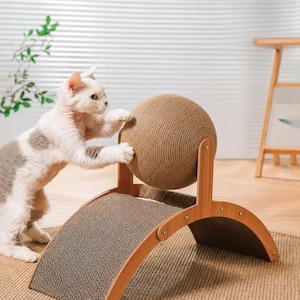 May include: A cat scratching toy featuring a large, round, tan sisal rope ball mounted on a curved, light-coloured wooden frame with a textured, corrugated cardboard scratching surface underneath the ball for feline claw maintenance.