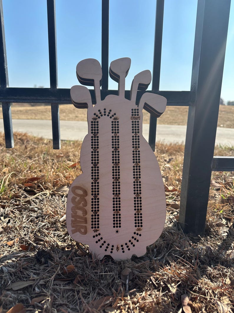 K&ouml;nnte beinhalten: Ein h&ouml;lzernes Cribbage-Brett in Form einer Golftasche mit Golfschl&auml;gern oben. Das Brett hat drei Bahnen f&uuml;r die Stifte und der Name "OSCAR" ist auf der Vorderseite eingraviert. Das Brett ist aus hellem Holz gefertigt.