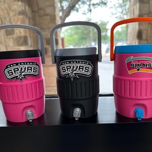May include: Three San Antonio Spurs water coolers in pink, black, and pink with blue accents. Each cooler has a handle and the Spurs logo. The coolers are cylindrical with a spigot at the bottom.