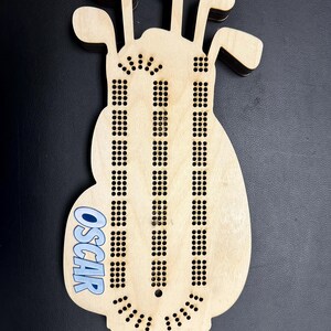 K&ouml;nnte beinhalten: Ein h&ouml;lzernes Cribbage-Brett in Form einer Golftasche mit Aussparungen f&uuml;r Golfschl&auml;ger oben. Das Brett hat Reihen von L&ouml;chern f&uuml;r die Stifte und den Namen "OSCAR" in Blau. Das Brett ist aus hellem Holz gefertigt.