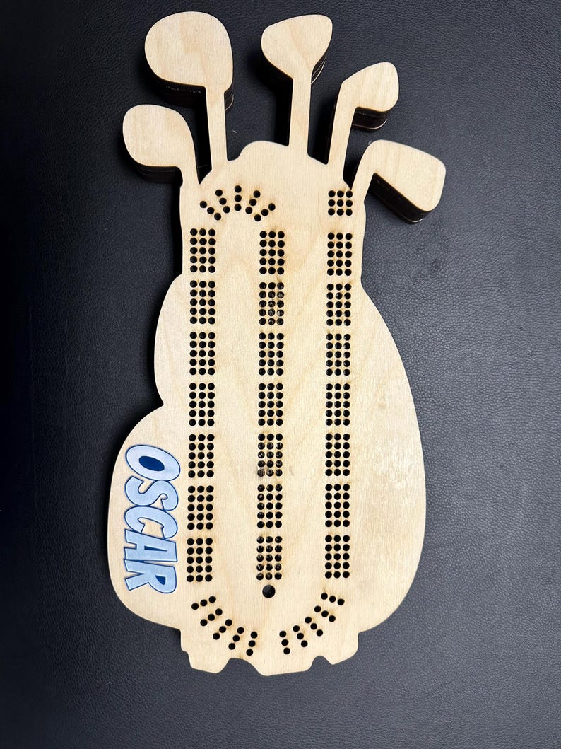 K&ouml;nnte beinhalten: Ein h&ouml;lzernes Cribbage-Brett in Form einer Golftasche mit Aussparungen f&uuml;r Golfschl&auml;ger oben. Das Brett hat Reihen von L&ouml;chern f&uuml;r die Stifte und den Namen "OSCAR" in Blau. Das Brett ist aus hellem Holz gefertigt.