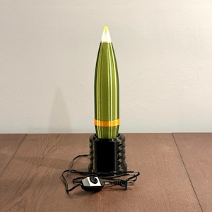 May include: A green and yellow lamp shaped like a bullet. The lamp has a black base and a yellow tip. An orange band encircles the lamp's body. The lamp is plugged into an electrical outlet and has an on/off switch.