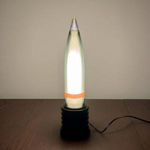 May include: A modern table lamp with a unique design. The lamp features a tall, bullet-shaped shade with a bright, internal light. The shade is supported by a black, cylindrical base. An orange band encircles the shade. The lamp is plugged into a dark wooden surface.