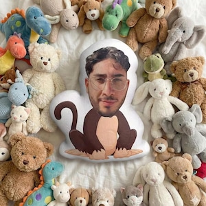 Peut inclure: Une collection de peluches, dont des ours en peluche, des dinosaures, des éléphants et des lapins, disposées autour d'un coussin en forme de singe avec le visage d'une personne. Les jouets sont de couleurs variées, créant une scène ludique et fantaisiste.