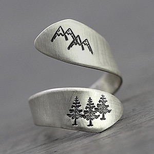 Puede incluir: Anillo envolvente plateado con diseños grabados de montañas y árboles. El anillo tiene un acabado de metal cepillado y un diseño abierto único. El diseño de la montaña está en la parte superior y los árboles en la inferior.