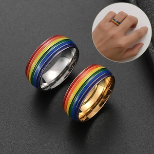 May include: Two rings with a rainbow design. One ring is silver, and the other is gold. The rings have a band of red, orange, yellow, green, blue, and purple. A close-up shows a ring on a finger.