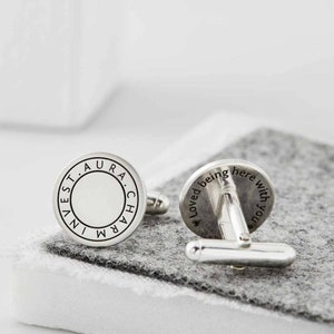 May include: A pair of silver cufflinks with engraved text. One cufflink says "IN. VEST. AURA. CHARM" and the other says "Loved being here with you".