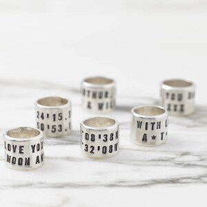 Puede incluir: Seis anillos de plata con diferentes textos grabados. El texto incluye fechas, coordenadas y frases como "Love You Moon And" y "With Love A*T".