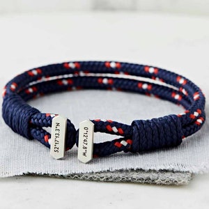 May include: A navy blue and red rope bracelet with two silver tags. The tags are engraved with the coordinates "52.1713.3'N" and "0.1247.8'W".
