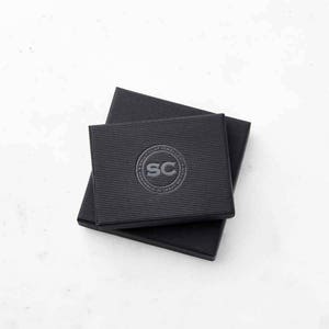 Puede incluir: Caja de cart&oacute;n negra con un logotipo que dice "Sally O'Cay Jewelry" y "Handmade in Greece" en un c&iacute;rculo alrededor de las letras "SC".