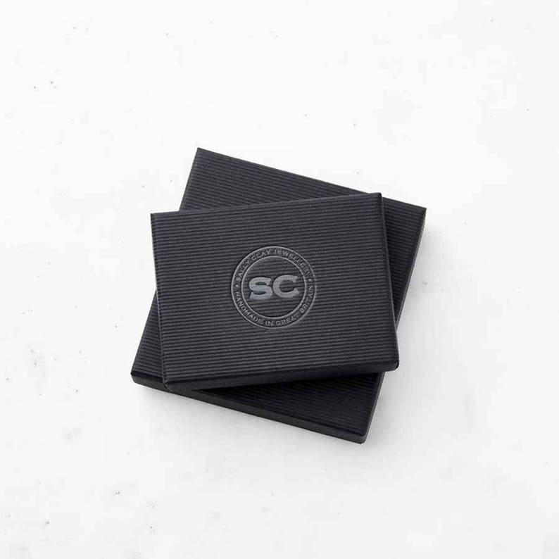 Puede incluir: Caja de cart&oacute;n negra con un logotipo que dice "Sally O'Cay Jewelry" y "Handmade in Greece" en un c&iacute;rculo alrededor de las letras "SC".