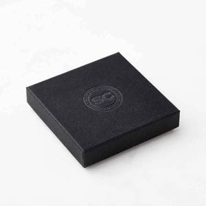 Peut inclure: Une bo&icirc;te carr&eacute;e noire avec une surface textur&eacute;e et un logo circulaire en relief sur le dessus. Le logo est de couleur argent&eacute;e et pr&eacute;sente les lettres "SC" &agrave; l'int&eacute;rieur d'un cercle.