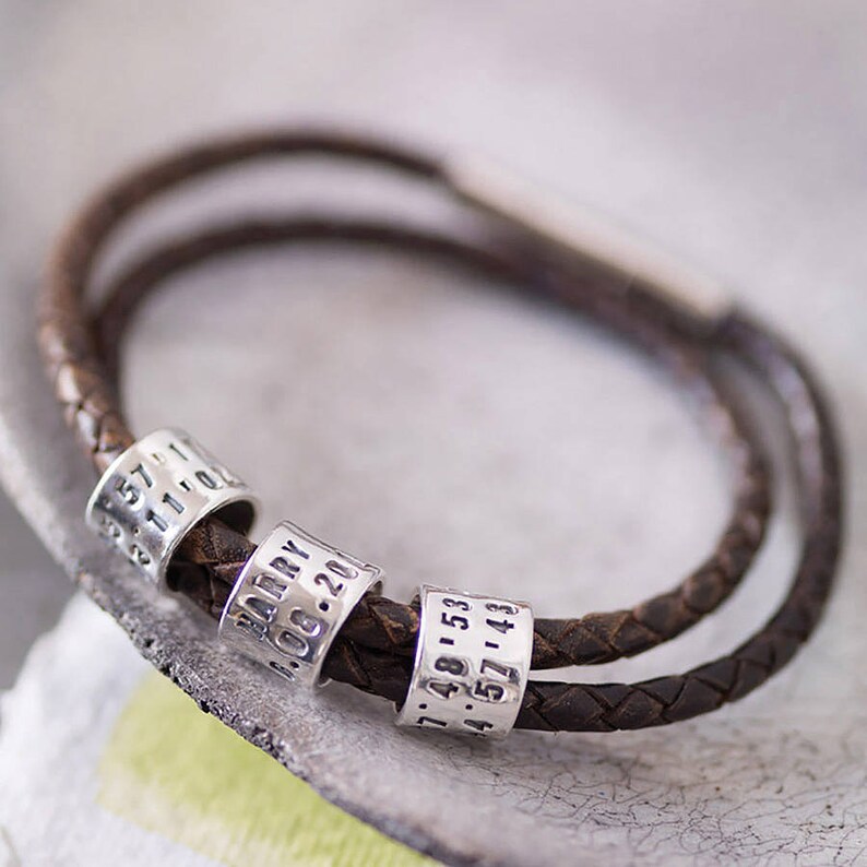 May include: A brown leather bracelet with three silver beads. The beads are engraved with the text "Harry", "06.20", and "1.48.153 4.57.143".