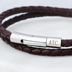 May include: A close-up of a brown leather bracelet with a braided design. The bracelet has a silver clasp with the initials "ATC" engraved. The bracelet is made of two strands of braided leather.