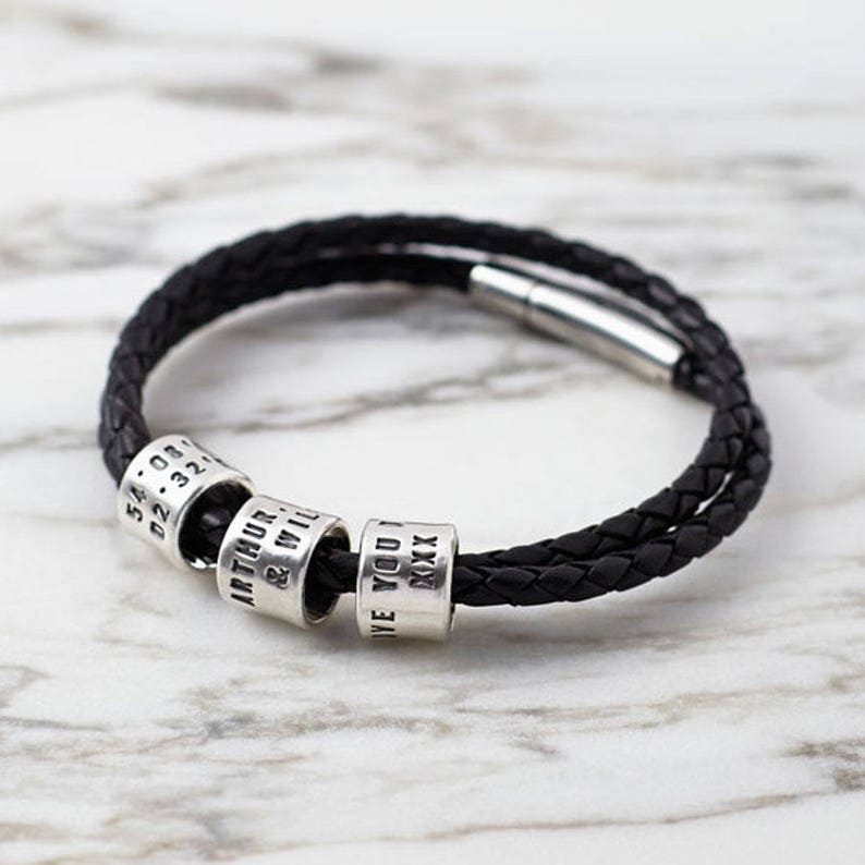 Puede incluir: Un brazalete de cuero negro con un dise&ntilde;o trenzado y cuatro cuentas de plata. Las cuentas est&aacute;n grabadas con el texto "54.08.02.32", "ARTHUR & WILL", "LOVE YOU" y "XXXX".