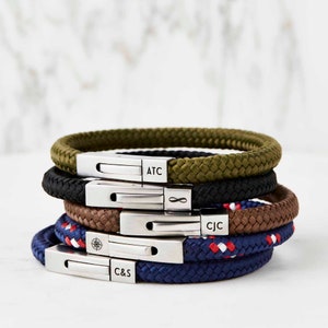 Op de afbeelding: Een stapel van vijf gepersonaliseerde touw armbanden met zilveren sluitingen. De armbanden zijn in verschillende kleuren, waaronder groen, zwart, bruin, blauw en rood. De sluitingen zijn gegraveerd met de initialen ATC, &infin;, CJC en C&S.