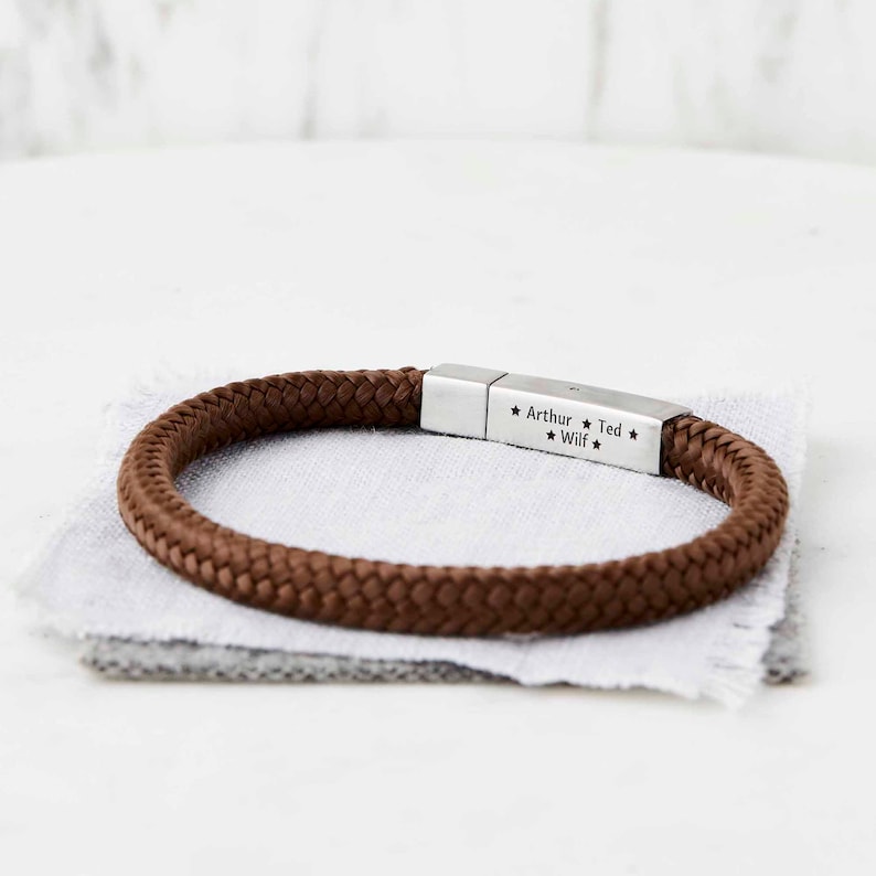 Op de afbeelding: Een bruin gevlochten touw armband met een zilveren metalen sluiting gegraveerd met de namen "Arthur", "Ted" en "Wilf" gescheiden door sterren.