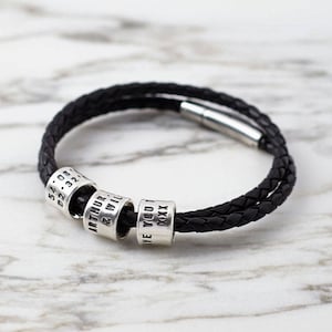 May include: A black leather bracelet with a braided design and four silver beads. The beads are engraved with the text "54.08.02.32", "ARTHUR & WILL", "LOVE YOU", and "XXXX".