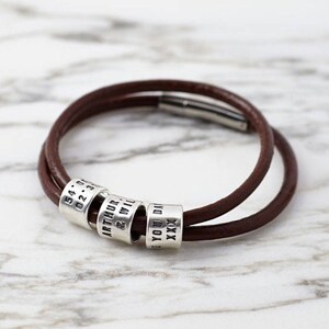 May include: A brown leather bracelet with four silver beads. The beads are engraved with the text "54.07.02.31", "ARTHUR & WIL", "YOU DA", and "XXX".