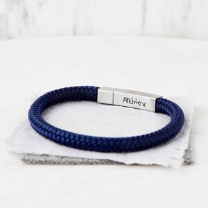 Pode incluir: Uma pulseira trançada azul marinho com um fecho prateado gravado com o nome "Archiex".