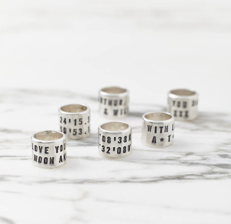 Puede incluir: Seis anillos de plata con diferentes textos grabados. El texto incluye fechas, coordenadas y frases como "Love You Moon And" y "With Love A*T".