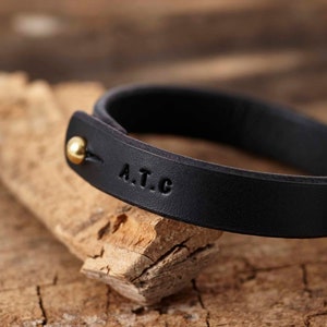 Peut inclure: Un bracelet en cuir noir avec un fermoir en or et les initiales "A.T.C." grav&eacute;es sur la bande.