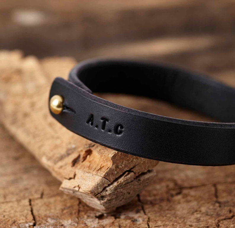 Peut inclure: Un bracelet en cuir noir avec un fermoir en or et les initiales "A.T.C." grav&eacute;es sur la bande.