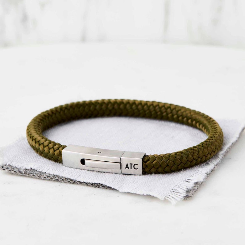 Op de afbeelding: Een groene gevlochten touw armband met een zilveren sluiting gegraveerd met de letters "ATC".