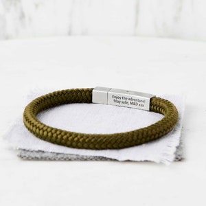 Op de afbeelding: Een groene touw armband met een zilveren metalen sluiting. De sluiting is gegraveerd met de tekst "Enjoy the adventure! Stay safe, M&D xxx".